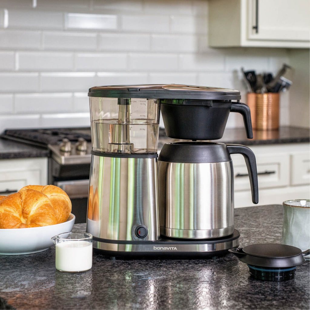 Una macchina Bonavita su un piano cucina in noce all'alba accanto a tazze di ceramica e un sacchetto di chicchi di caffè freschi